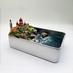 Könnte beinhalten: Miniatur-Diorama einer Küstenlandschaft mit einem detaillierten Gebäude mit roten und orangen Kuppeln und Türmen. Die Szene befindet sich in einem silbernen Metallbehälter mit blauem Ozean und felsigen Klippen.
