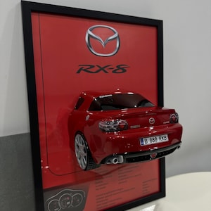 May include: Framed artwork featuring a red Mazda RX-8 sports car model. The car is mounted on a red background with the Mazda logo and RX-8 text. The frame is black.