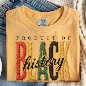 Op de afbeelding: Mosterdgeel T-shirt met de geborduurde tekst "PRODUCT OF BLACK history". De letters zijn rood, geel, groen en zwart. Het shirt is opgevouwen op een blauwe spijkerbroek en een witte sneaker.