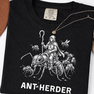 Puede incluir: Camiseta negra con un gráfico blanco detallado de una figura encapuchada montada en una hormiga gigante, con el texto "ANT-HERDER" debajo. El diseño incluye varias hormigas más pequeñas y una abeja. Se ve un collar dorado y un sombrero marrón.