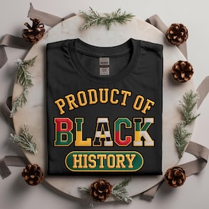 May include: Black t-shirt featuring the text "PRODUCT OF BLACK HISTORY" in a collegiate font. The design uses red, yellow, green, and black. The shirt is presented on a decorative surface with pine cones and sprigs of greenery.
