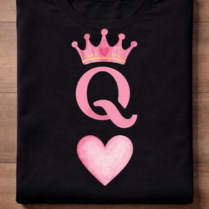 Peut inclure: T-shirt noir avec une couronne rose, la lettre "Q" et un cœur. La couronne a une bordure dorée et un cœur rose au centre. Le "Q" est en gras, sans empattement. Le cœur est de style aquarelle.