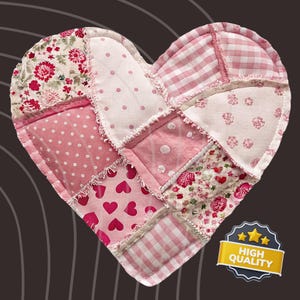 Peut inclure: Un objet en patchwork en forme de cœur, composé de divers tissus roses. Le motif comprend des motifs floraux, à pois, à carreaux et en forme de cœur. Les bords ont une finition effilochée. Un badge "HIGH QUALITY" est en bas à droite.