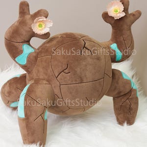 May include: A brown plush toy with a cracked, spherical body and four stubby legs. It has two arms with pink flower accents and turquoise markings. The toy is on a white, fluffy surface.