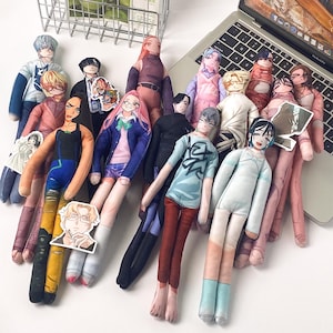 May include: A collection of flat, fabric-printed plush dolls featuring various anime-style characters in diverse outfits, including shirts, sweaters, and pants. Several small, separate character stickers are scattered among the plush figures.