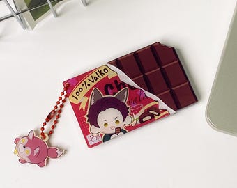 May include: A rectangular acrylic charm shaped like a chocolate bar with a red wrapper featuring the text 100% Valko and an illustration of a character with animal ears. A small red ball chain attaches a separate pink fox-like character charm.