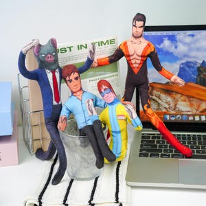 May include: A collection of handmade fabric dolls, each with unique designs. One doll is dressed in a blue suit with a pig-like mask, another in a superhero costume, and others in casual attire. The dolls are arranged near a laptop and a newspaper.