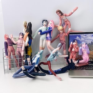 May include: A collection of flat, printed fabric dolls featuring various anime-style characters in diverse outfits, including pink, blue, and dark clothing. The dolls have elongated limbs and are arranged in a group, some standing and some reclining.