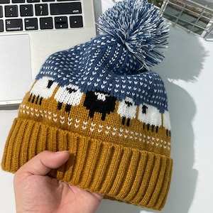 Könnte beinhalten: Eine Strickmütze mit senfgelbem Rippbündchen und einer blau-weiß gesprenkelten Krone. Die Mütze zeigt ein sich wiederholendes Muster aus weißen und schwarzen Schafen in der Mitte und ist mit einem großen blau-weißen Bommel versehen.