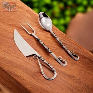 May include: A handcrafted, three-piece flatware set. The set includes a knife, fork, and spoon. Each piece has a twisted handle design and a polished, silver-colored finish. The set is displayed on a wooden surface.