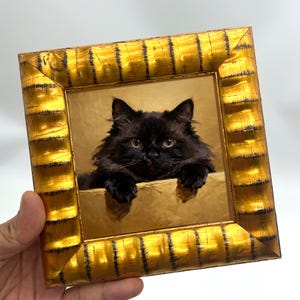 Puede incluir: Pintura enmarcada de un gato negro con pelaje esponjoso, asomándose por encima de una repisa dorada. Los ojos del gato son visibles y el marco es de color dorado texturizado. La obra de arte está sujeta por una mano.