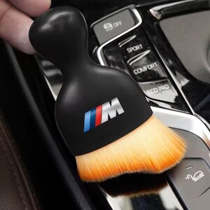 K&ouml;nnte beinhalten: Eine schwarze Autoreinigungsb&uuml;rste mit dem BMW-Logo in Blau, Rot und Wei&szlig;. Die B&uuml;rste hat einen abgerundeten Griff und einen Kopf aus orangefarbenen und hellbraunen Borsten. Sie ist f&uuml;r die Innenreinigung eines Fahrzeugs konzipiert.