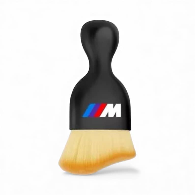 K&ouml;nnte beinhalten: Ein schwarzer Make-up-Pinsel mit gebogenem Griff und einem Logo mit blauen, roten und wei&szlig;en Streifen und dem Buchstaben "M". Der Pinselkopf ist hellbraun.