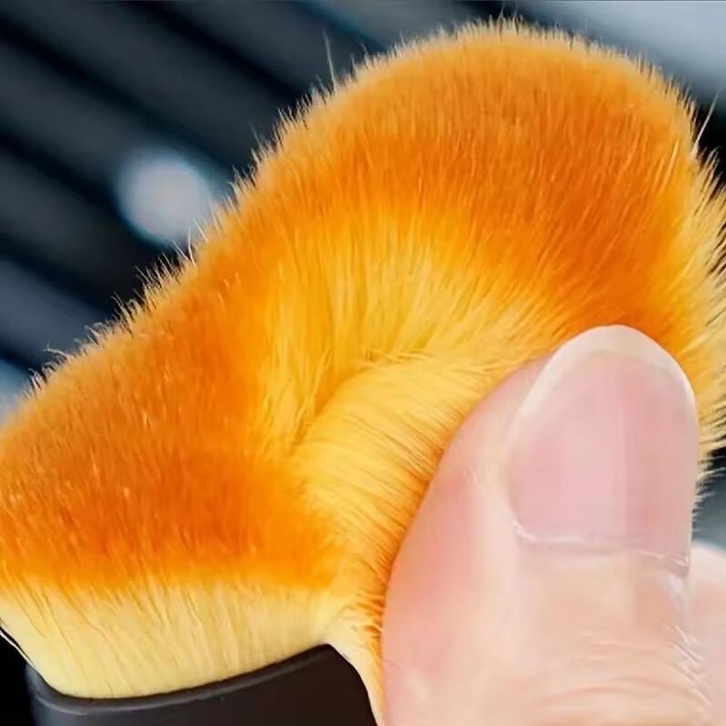 K&ouml;nnte beinhalten: Nahaufnahme eines Make-up-Pinsels mit dichtem, flauschigem Kopf. Der Pinselkopf hat einen Farbverlauf von Orange und Gelb mit einem weichen, strukturierten Aussehen. Der Griff ist schwarz, und ein Finger ber&uuml;hrt den Pinsel.