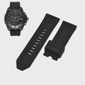 May include: A black wristwatch with a black rubber strap and a separate black rubber watch strap. The watch face has multiple dials and a date window. The watch and straps are set against a light gray background.