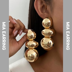 Puede incluir: Pendientes dorados llamativos con un diseño de bolas apiladas. Los pendientes tienen un acabado brillante y reflejan el entorno. El texto "MIA EARRING" está impreso verticalmente en el lado izquierdo de la imagen.