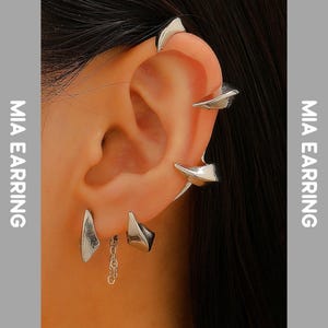 Könnte beinhalten: Silberfarbenes Ohr-Cuff- und Ohrring-Set. Das Ohr-Cuff hat eine Reihe von spitzen, kantigen Designs, die sich entlang des Ohrs krümmen. Die Ohrringe umfassen zwei dreieckige Ohrstecker und eine hängende Kette. Der Text "MIA EARRING" ist auf dem Bild zu sehen.