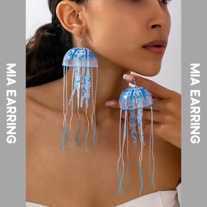 Può includere: Un paio di orecchini a forma di medusa blu e bianchi. Gli orecchini hanno una cupola traslucida con macchie blu e lunghi tentacoli fluenti. Il testo "MIA EARRING" è visibile.