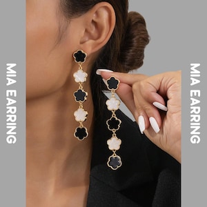 Peut inclure: Longues boucles d'oreilles pendantes avec un motif floral. Les boucles d'oreilles alternent des pendentifs en forme de fleurs noires et blanches, reliés par des chaînes dorées. Le texte "MIA EARRING" est imprimé verticalement.
