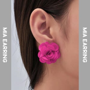 Puede incluir: Primer plano de un pendiente en forma de flor rosa brillante. El pendiente se lleva en una oreja, con el texto "MIA EARRING" en los lados izquierdo y derecho de la imagen. Un accesorio llamativo.