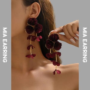 Op de afbeelding: Bordeaux bloemvormige oorhangers met goudkleurige accenten. De oorbellen hebben gelaagde stoffen bloemen en delicate gouden stelen. De tekst "MIA EARRING" staat verticaal op de linker- en rechterkant van de afbeelding.