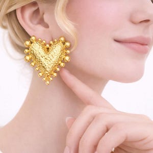 Puede incluir: Pendientes dorados en forma de corazón. Los pendientes tienen una superficie texturizada y están adornados con pequeños adornos redondos en los bordes. Los pendientes se llevan en la oreja de una persona.