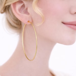 Puede incluir: Pendientes de aro dorados. Los pendientes son grandes, de forma ovalada y cuelgan de la oreja. La imagen muestra un primer plano de los pendientes y parte de la cara y el hombro de la persona. Los pendientes tienen un diseño sencillo y elegante.