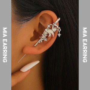 Könnte beinhalten: Silberner Ohrklemme mit einem Schwert-, Stern- und Mond-Design. Die Manschette umschließt das Ohr und verfügt über einen baumelnden Sternanhänger und eine weiße Perle. Der Text "MIA EARRING" befindet sich auf der linken und rechten Seite des Bildes.