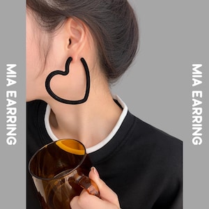 Puede incluir: Pendientes de aro negros en forma de corazón. Los pendientes se llevan en la oreja. La imagen también muestra una camisa negra con cuello blanco y el texto "MIA EARRING" en el lateral.