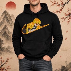 Op de afbeelding: Zwarte hoodie met een geel en oranje grafisch ontwerp. Het ontwerp toont een anime-personage in een swoosh-logo. De hoodie heeft een voorzak en een capuchon met trekkoord.