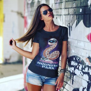 Könnte beinhalten: Schwarzes T-Shirt mit einer Comic-Schlange, die eine VR-Brille trägt. Die Schlange ist hellbraun mit weißem Bauch. Das Shirt trägt den Text "Longitano Hognose Park" in einem Retro-Schriftzug. Das Shirt wird mit blauen Jeansshorts getragen.