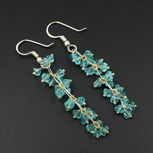 Puede incluir: Un par de pendientes colgantes con ganchos plateados y delicados hilos de cuentas de cristal azul claro. Las cuentas están agrupadas, creando un diseño floral. Los pendientes se presentan sobre un fondo oscuro.