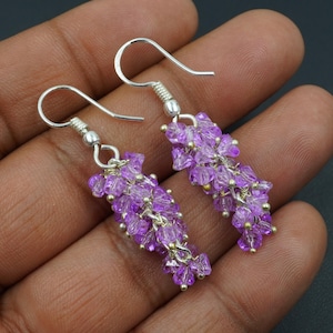 Puede incluir: Un par de pendientes colgantes con racimos de pequeñas cuentas moradas facetadas. Las cuentas están ensartadas y unidas a ganchos para las orejas de color plateado. Los pendientes tienen un diseño delicado y en cascada.