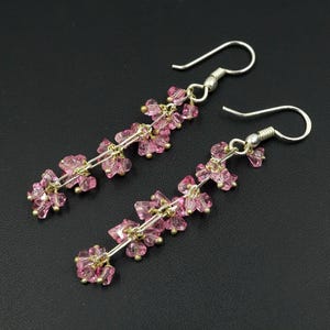 Puede incluir: Un par de pendientes colgantes con un diseño floral. Cada pendiente tiene un gancho plateado y una serie de cuentas rosas facetadas ensartadas en un alambre plateado. Las cuentas están agrupadas para parecer pétalos de flores.