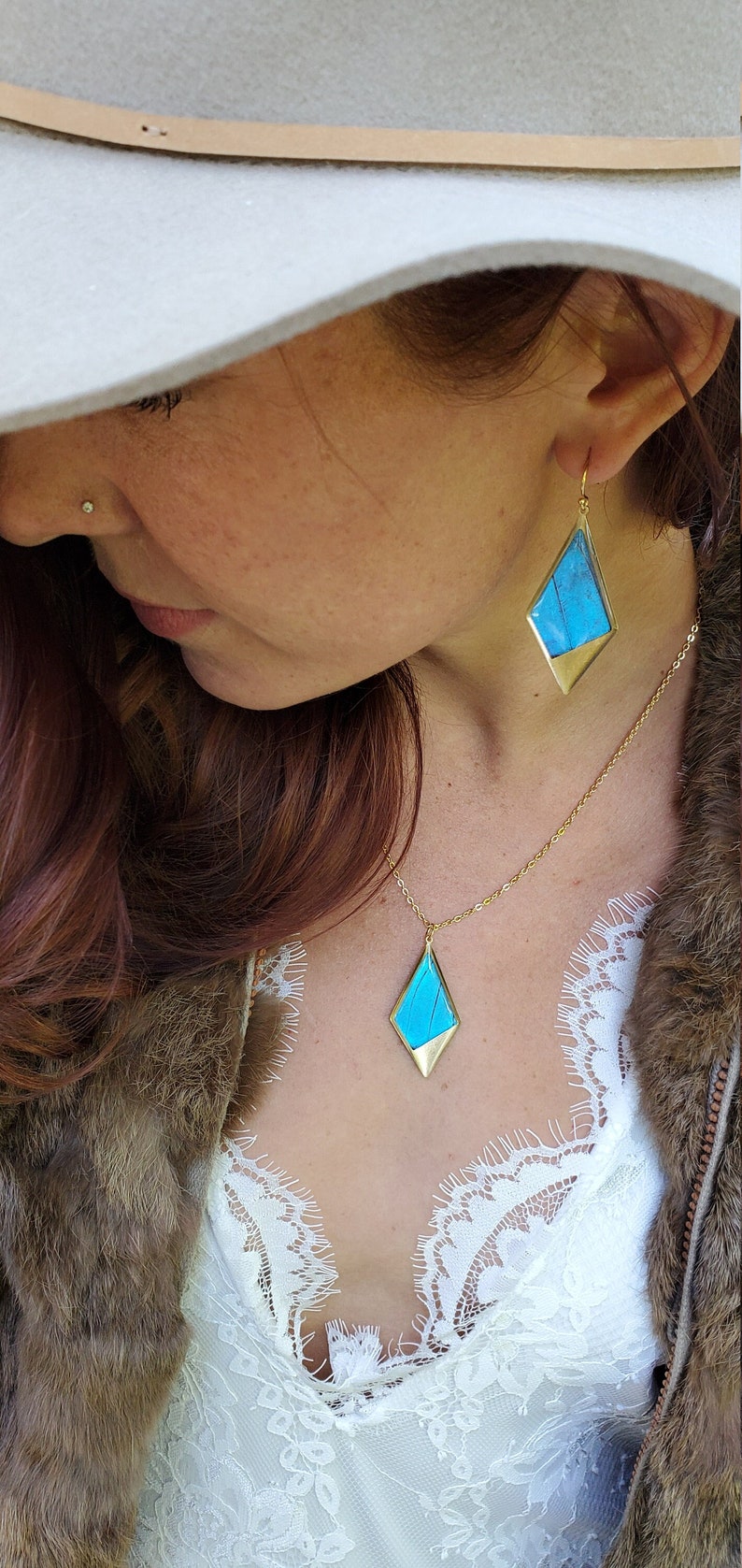 May include: A woman wearing a white hat and a white lace top with a gold necklace and earrings featuring blue butterfly wings. The necklace and earrings have a diamond shape.