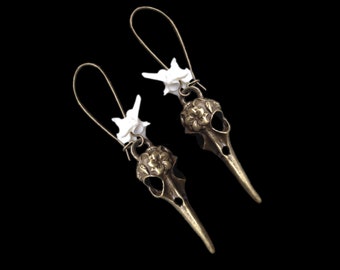 Puede incluir: Un par de pendientes de bronce en forma de cráneo de pájaro con detalles en hueso blanco. Los pendientes presentan un diseño floral en el cráneo y están suspendidos de un simple bucle de alambre.
