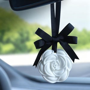 Könnte beinhalten: Ein weißes, rosenförmiges Ornament, das an einem schwarzen Band hängt. Das Band ist oben zu einer dekorativen Schleife gebunden und hält das blumenförmige Stück. Dieses hängende Accessoire ist zur Befestigung am Rückspiegel gedacht.