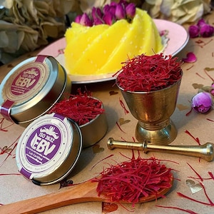 Puede incluir: Arreglo con hebras de azafrán, un mortero y una maja dorados, y pequeñas latas de azafrán. Una cuchara de madera contiene hebras de azafrán. Un postre amarillo está sobre un plato rosa, y capullos de rosa rosas.