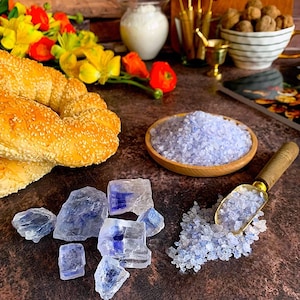 May include: A braided loaf of bread topped with sesame seeds, a small wooden bowl filled with light purple coarse salt, several translucent blue-tinted rock crystals, and a wooden scoop containing more of the same light purple salt.