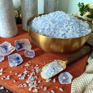 Puede incluir: Un cuenco de latón lleno de sales de baño de color azul claro, acompañado de una pala de metal a juego con mango de madera. Varios trozos grandes y translúcidos similares a cristales azules están esparcidos por la superficie.