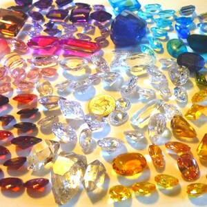 May include: A large collection of loose faceted gemstones in various shapes and colors, including clear, yellow, orange, red, purple, and blue stones. A silver-colored United States dime is positioned near the center for scale reference.