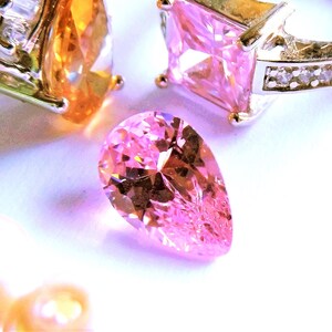 a pink faceted pear cut CZ gemstone sits near finished jewelry pieces, ready to be placed in your setting.