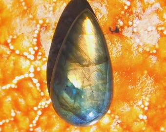 May include: A teardrop-shaped labradorite gemstone with iridescent blue, green, and gold hues. The polished stone is set against an orange background with a white dotted pattern. The gemstone has a smooth, reflective surface.