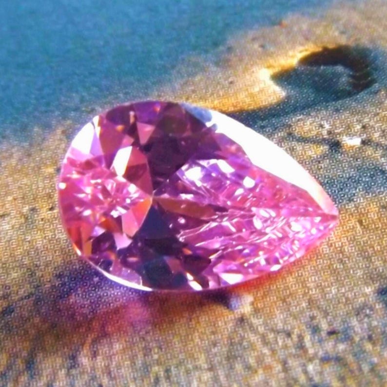 Vivid pink pear cut CZ gem on its side on a sandy background with a footprint behind it, reflecting light and shadow beneath it.