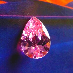 A pink CZ teardrop cut, faceted gemstone with a blue and mountainous background