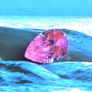 a pink CZ pear cut faceted stone seems to be balancing on a wet blue rocky surface with a blue watery background