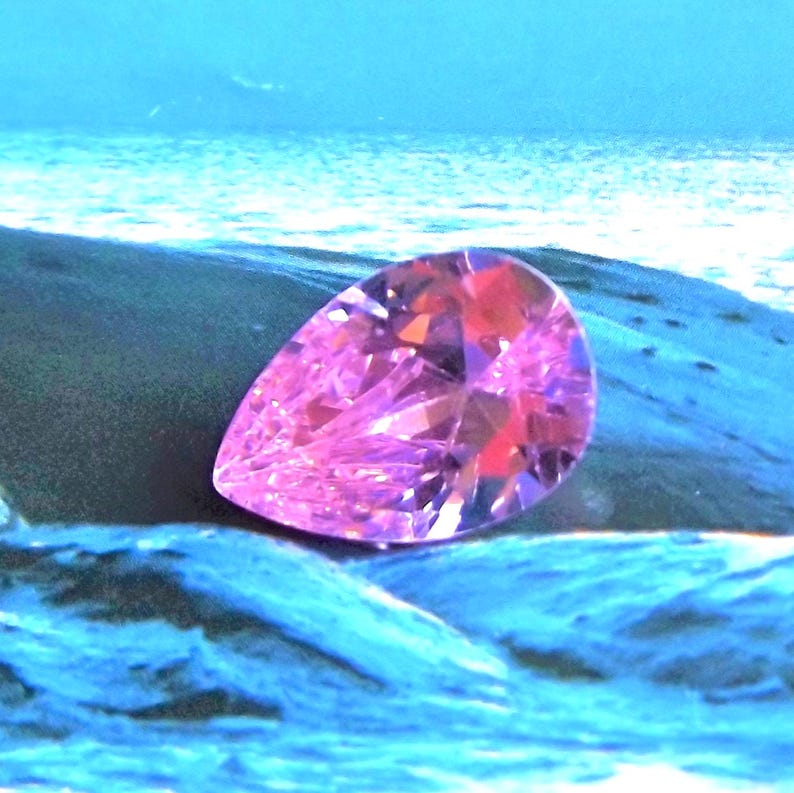 a pink CZ pear cut faceted stone seems to be balancing on a wet blue rocky surface with a blue watery background