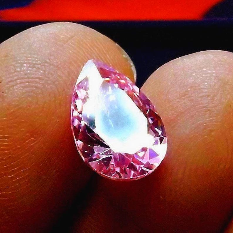A faceted pink Cubic Zirconia gemstone balanced on two fingers to show size/scale.