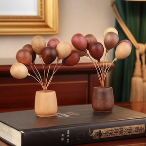 Puede incluir: Dos ramos de globos de madera en pequeños jarrones. Los globos son de varios tonos de marrón y beige, con finos tallos dorados. Los jarrones son de diferentes tonos de marrón. Los ramos están sobre un libro oscuro.