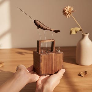 May include: A wooden narwhal sculpture with a long tusk is suspended above a wooden box. The sculpture is supported by thin metal rods. A small, beige vase with a dried flower sits to the right. The sculpture is held by a person.